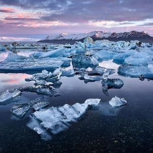 Iceberg e blocchi di ghiaccio galleggiano in una laguna calma al tramonto, con montagne innevate in lontananza sotto un cielo rosa e viola.