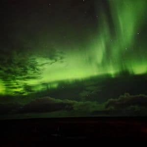 Vivide luci verdi dell'aurora boreale illuminano il cielo notturno sopra un paesaggio scuro con nuvole sparse e stelle visibili.