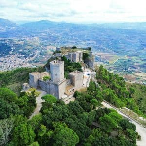 Una vista aerea di un castello storico in pietra, situato su una montagna boscosa, che domina un'ampia valle e una città lontana.