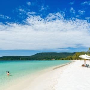 Una vasta distesa di spiaggia di sabbia bianca incontra acque limpide e turchesi, con una grande collina verde in lontananza sotto un cielo azzurro.