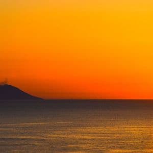 Eine Vulkaninsel zeichnet sich vor einem leuchtend orangefarbenen Sonnenuntergang über dem Meer ab, aus ihrem Gipfel steigt Rauch auf.