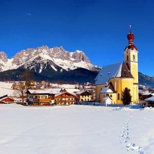 Eine gelbe Kirche in einem schneebedeckten Dorf mit Fußspuren im Vordergrund, vor einer Kulisse von Bergen unter einem klaren blauen Himmel.