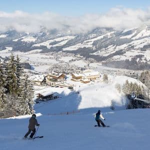 Ein Skifahrer und ein Snowboarder von einer WeRoad Gruppenreise fahren einen sonnigen Hang oberhalb eines schneebedeckten Bergdorfs hinab.