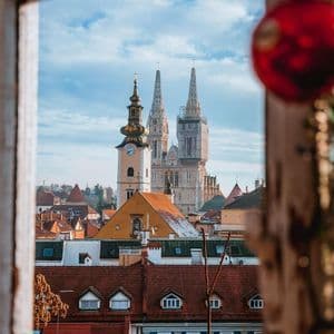 A cityscape with a cathedral and a clock tower seen through a window, with a red Christmas ornament blurred in the foreground.