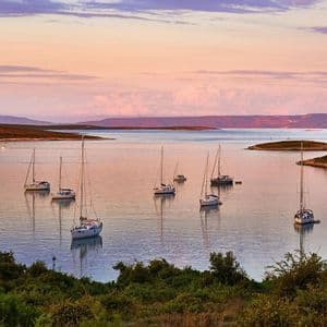 Several sailboats are anchored in a tranquil bay surrounded by islands under a pastel-colored sunset sky.
