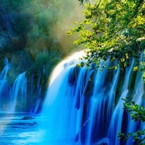 A waterfall with a silky motion-blur effect tumbles into a pool, framed by lush green trees with sunlight filtering through the leaves.