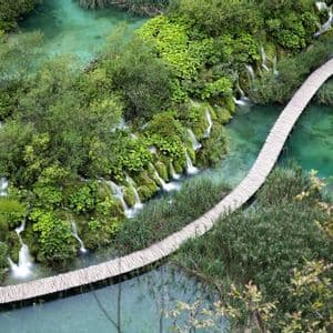 An aerial view of a wooden boardwalk winding over a turquoise lake, surrounded by lush green foliage and small waterfalls.