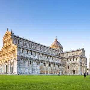 Una catedral ornamentada y un campanario inclinado sobre un gran césped verde bajo un cielo azul claro durante el día.