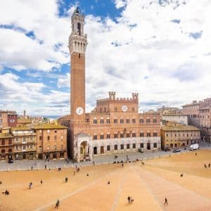 Vista elevada de una vasta y histórica plaza europea, con un alto campanario de ladrillo y personas en la plaza.