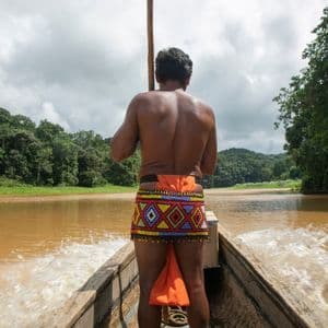 Un uomo in abiti tradizionali con perline, visto da dietro, guida una barca di legno lungo un ampio fiume costeggiato da una fitta giungla verde.
