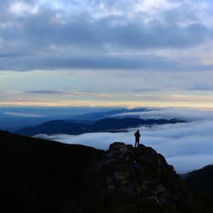 Una persona si staglia in silhouette su una vetta rocciosa di montagna, ammirando un vasto mare di nuvole e montagne lontane.