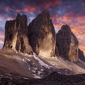 Drei zerklüftete Berggipfel mit Schneefeldern an ihren Hängen, beleuchtet von einem dramatischen und farbenprächtigen Sonnenuntergang.