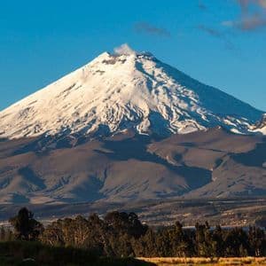 Un gran volcán cubierto de nieve emite una columna de humo desde su cima, dominando un paisaje de colinas y árboles bajo un cielo azul claro.