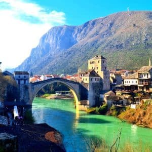 Eine alte Steinbogenbrücke überspannt einen lebhaft türkisfarbenen Fluss, der durch eine historische Stadt fließt, mit einem hohen Berg im Hintergrund.