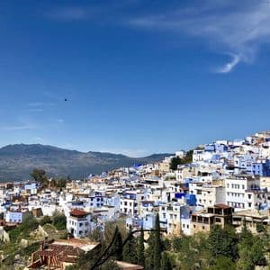 A panoramic view of a city with blue and white buildings on a hillside, with mountains in the background under a clear sky.