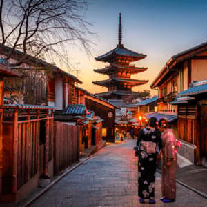 Two women in traditional kimonos stand on a historic street facing a Japanese pagoda at sunset.