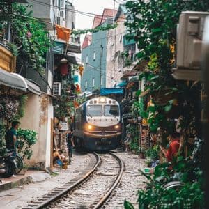 Un treno con il faro acceso viaggia lungo i binari attraverso una stretta via residenziale fiancheggiata da edifici e vegetazione lussureggiante.