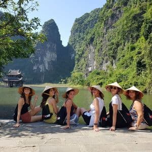 A WeRoad group trip of six women wearing conical hats sitting on a stone pier by a lake, with a pagoda and green karst mountains in the background.