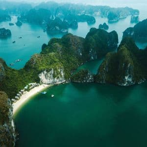 Aerial view of numerous green-topped limestone islands rising from turquoise water, with a secluded white sand beach and bungalows.