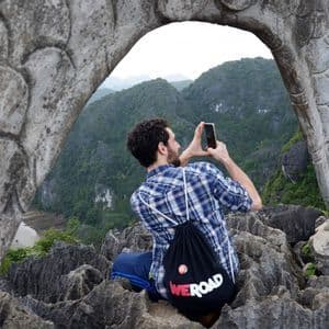 Un hombre con una mochila WeRoad se sienta en un mirador rocoso, tomando una foto de las montañas a través de un arco de dragón de piedra.