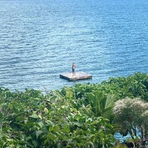 Un homme en short se tient sur un radeau en bois flottant sur un grand lac, entouré de montagnes vertes et d'une végétation luxuriante.