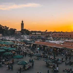 A high-angle view of a bustling city square filled with market stalls and people at sunset, with a prominent tower in the distance.
