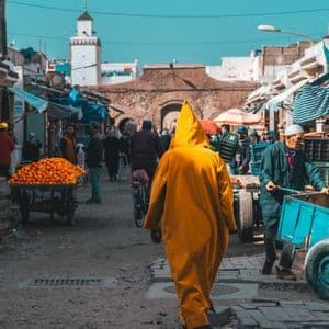 A person wearing a yellow hooded robe walks down a busy market street with a cart of oranges and a minaret in the distance.