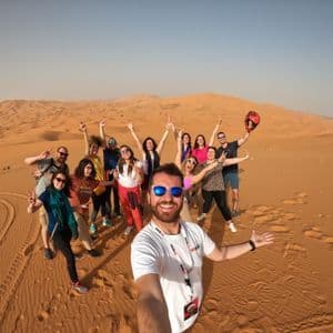 A man takes a selfie with a WeRoad group trip as they pose and smile on large sand dunes in a desert.