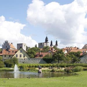 Eine historische Stadt mit Steintürmen und rot gedeckten Häusern, gesehen von einem grünen Park mit Springbrunnen in einem Teich.