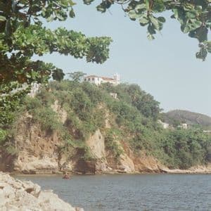 Ein weißes Gebäude auf einer üppigen, felsigen Klippe mit Blick auf das Wasser, gesehen hinter Felsen und unter Baumästen.
