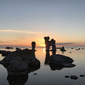 Silhouetten großer Felsformationen stehen im ruhigen Wasser, während die Sonne am Horizont untergeht.