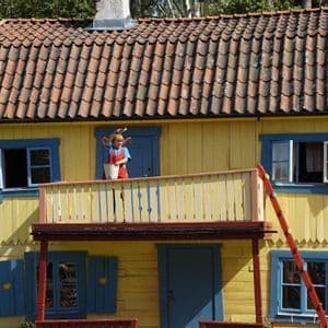 Eine Schaufensterpuppe von Pippi Langstrumpf steht auf dem Balkon eines gelben Hauses und hält einen weißen Eimer und einen Pinsel.