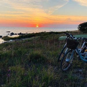 Zwei Fahrräder stehen an einer grasbewachsenen Küste mit Blick auf den Ozean, während die Sonne am Horizont untergeht.