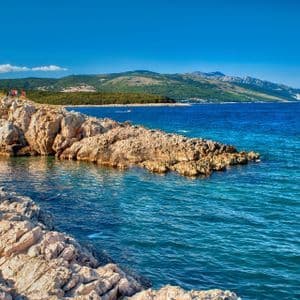 Una costa rocciosa si protende in un mare calmo e blu, con lontane colline verdi sotto un cielo sereno in una giornata di sole.