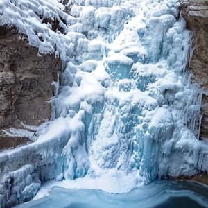 Una grande cascata ghiacciata e innevata scende da un canyon roccioso in una pozza d'acqua scura e gelida sottostante.