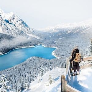 Un escursionista con uno zaino si trova su un punto panoramico innevato che domina una valle con un lago blu, foreste innevate e montagne.