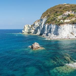 Vista dall'alto di una cala assolata con scogliere bianche e una collina verde, dove acque turchesi limpide lambiscono una piccola spiaggia con persone.