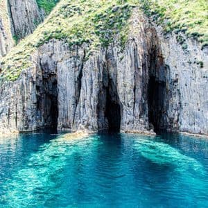 Diverse grotte marine scure alla base di una scogliera rocciosa con la cima erbosa, che si affacciano su acque limpide e turchesi.