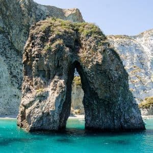 Un grande arco naturale di roccia si erge in acque turchesi, con una spiaggia e scogliere bianche sullo sfondo.