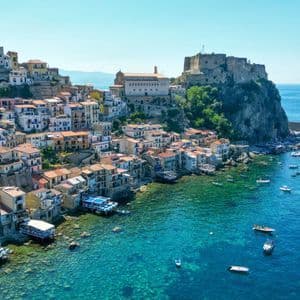 Una veduta aerea di una cittadina colorata costruita su una scogliera sul mare, con un castello che domina un porto pieno di barche in acque turchesi.