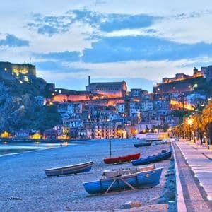 Barche riposano su una spiaggia sabbiosa accanto a un lungomare, con un paese illuminato sulla scogliera sotto un cielo crepuscolare.