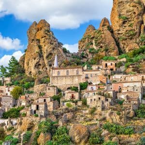 Un borgo storico con case in pietra e una chiesa incastonata alla base di formazioni rocciose frastagliate sotto un cielo parzialmente nuvoloso.