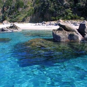 Acqua turchese limpida increspa grandi rocce davanti a una spiaggia isolata di sabbia bianca e una collina verdeggiante.