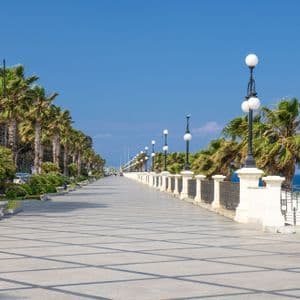 Un ampio lungomare piastrellato, costeggiato da alte palme e lampioni ornati, corre lungo il mare sotto un cielo azzurro e limpido.