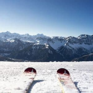 Una visuale in prima persona dalle punte degli sci su un pendio innevato, di fronte a un'ampia catena di montagne innevate sotto un cielo sereno.