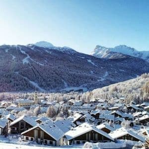 Una vista grandangolare di un villaggio alpino innevato con chalet incastonati ai piedi di montagne boscose in una giornata di sole.