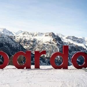 Una donna in tenuta da sci posa sugli sci accanto a grandi lettere rosse che formano la parola 'bardo' in un paesaggio montano innevato.