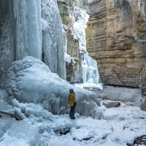Una persona con una giacca invernale gialla si trova in un canyon innevato, guardando verso enormi cascate ghiacciate aggrappate alle pareti rocciose.