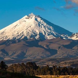 A large, snow-capped volcano emitting a plume of smoke, set against a blue sky above a valley with trees.