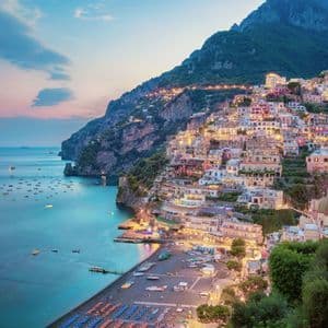 A colorful village built on a steep cliffside is illuminated at dusk, overlooking a calm bay filled with many boats.